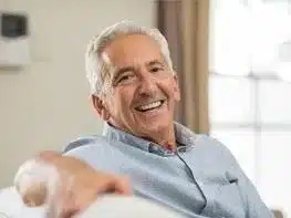 Smiling senior man in light blue shirt gesturing in bright dental office setting.