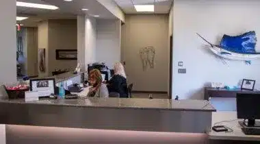 Dental receptionist at front desk in modern dental office with waiting area visible behind counter.
